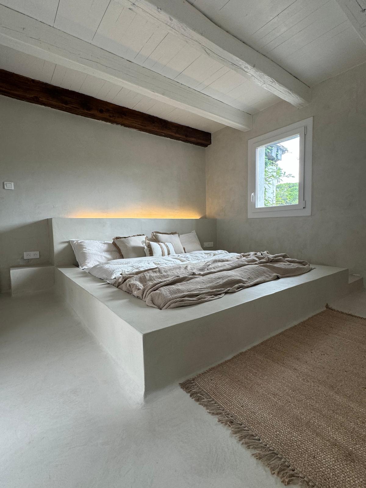 Chambre épurée, lit plateforme, murs blancs et poutres bois, lumière naturelle, style béton ciré élégant.