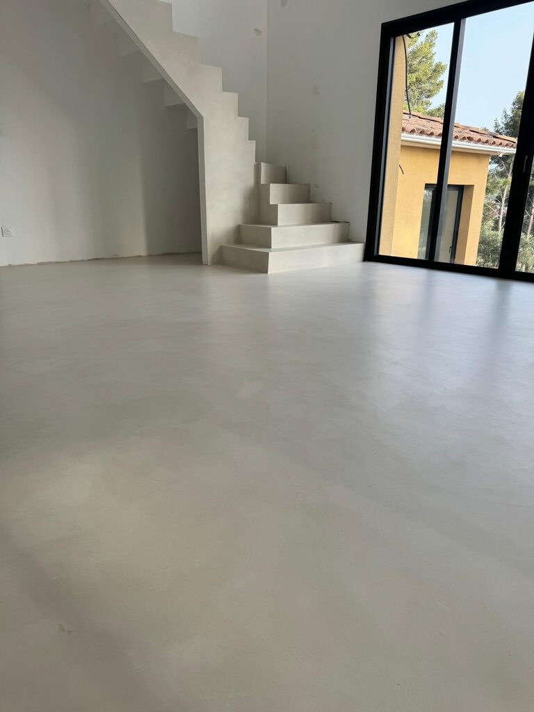 Intérieur épuré, sol béton ciré blanc, escalier angulaire, larges fenêtres et lumière naturelle.