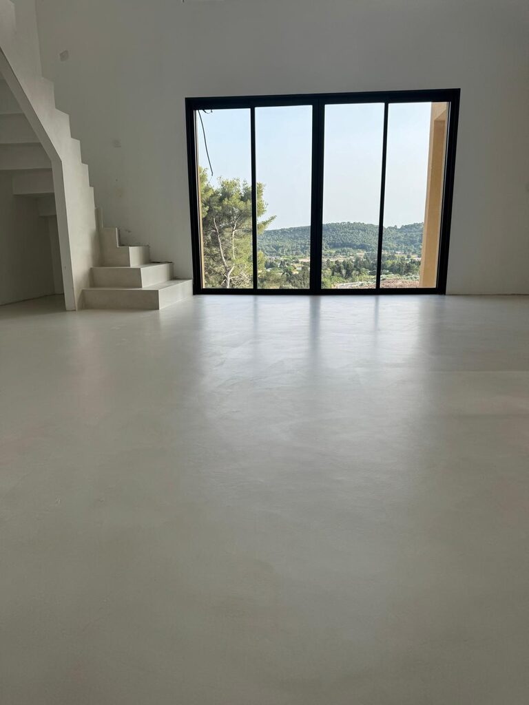 Espace minimaliste, sol blanc, escaliers, larges baies sur vue verdoyante et collines illuminées.