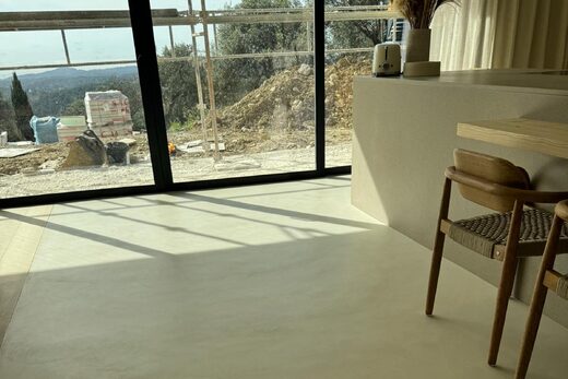 Espace épuré, sol clair, chaises bois, larges baies vitrées sur chantier et paysage.